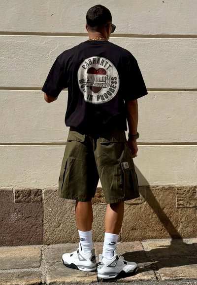 Camiseta negra con un estampado gráfico que presenta un camión y texto, combinada con pantalones cortos cargo de color verde oliva y zapatillas blancas.