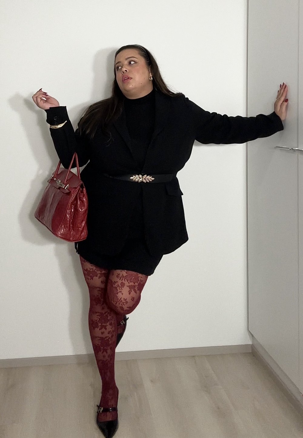 Femme en blazer noir et jupe, portant des collants rouges à motifs floraux, tenant un sac à main rouge, appuyée contre un mur blanc à l'intérieur.