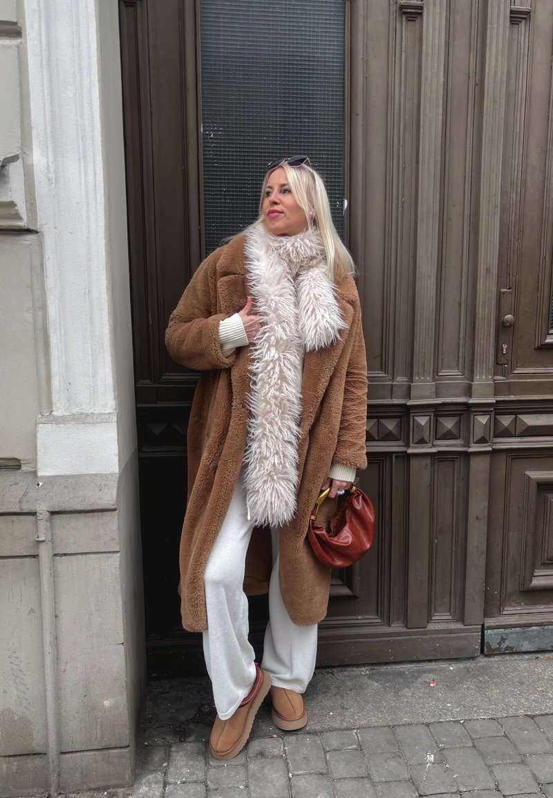 Frau in beigefarbenem Pelzmantel, weißen Hosen und Schal, die eine kleine braune Handtasche hält und auf dem Bürgersteig vor einer verzierten Holztür steht.