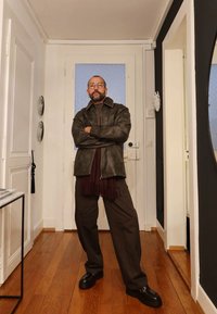 Brązowa skórzana kurtka, bordowy szalik i ciemne spodnie. Czarne buty z ozdobami. Stoi w dobrze oświetlonym wnętrzu z drewnianą podłogą.