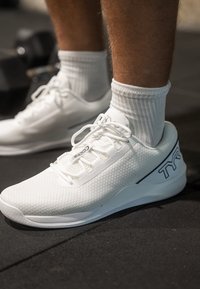 Weisse Sport Schuhe mit Mesh-Obermaterial, flacher Sohle und schwarzem Logo an der Seite, kombiniert mit gerippten weissen Socken, vor einem Fitnessboden.