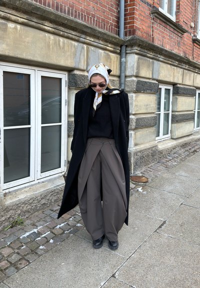 Mujer con abrigo negro, pantalones anchos grises, zapatos negros, gafas de sol y pañuelo blanco con estampado, de pie en la acera cerca de un edificio de piedra.