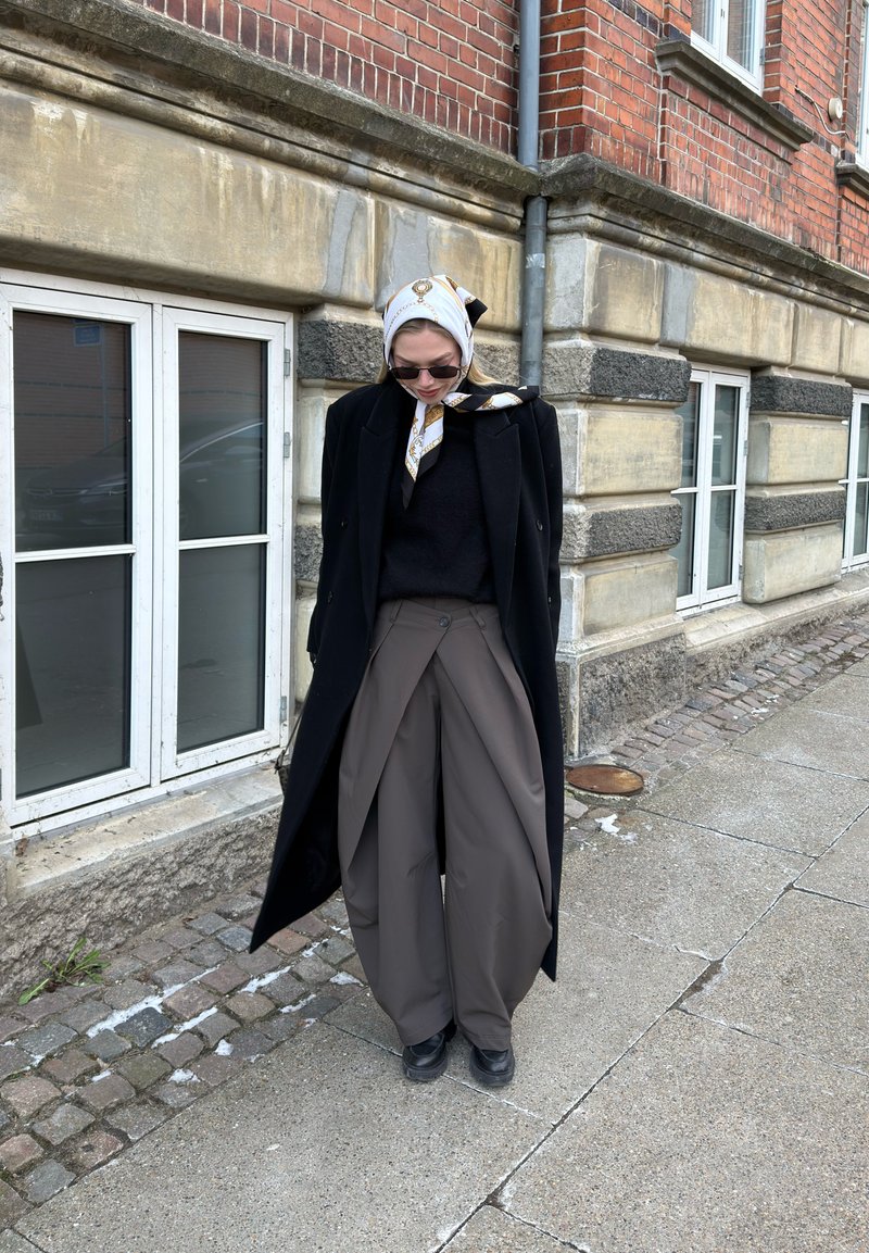 Femme portant un manteau noir, un pantalon gris large, des chaussures noires, des lunettes de soleil et un foulard blanc à motifs, debout sur le trottoir près d'un bâtiment en pierre.