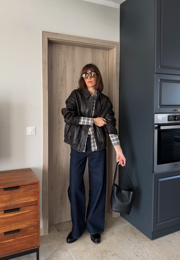 Naine, kes kannab päikeseprille, musta nahktagi, ruudulist särk ja laiade säärega teksaseid, hoides musta käekotti moodsa köögi ukseavas.