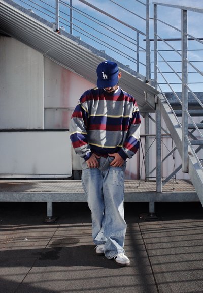 Jersey con un patrón de rayas en gris, rojo y amarillo; vaqueros azules holgados con las letras 'KS' bordadas; zapatillas blancas; gorra azul.