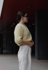 Žlutý žebrovaný pletený svetr s krátkými rukávy, nošený přes bílé kalhoty s vysokým pasem. Hladká textura, volný střih a moderní silueta.