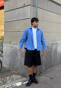 Camisa azul claro abotonada sobre una camiseta blanca, combinada con pantalones cortos negros y zapatos negros. De pie junto a una pared de piedra.
