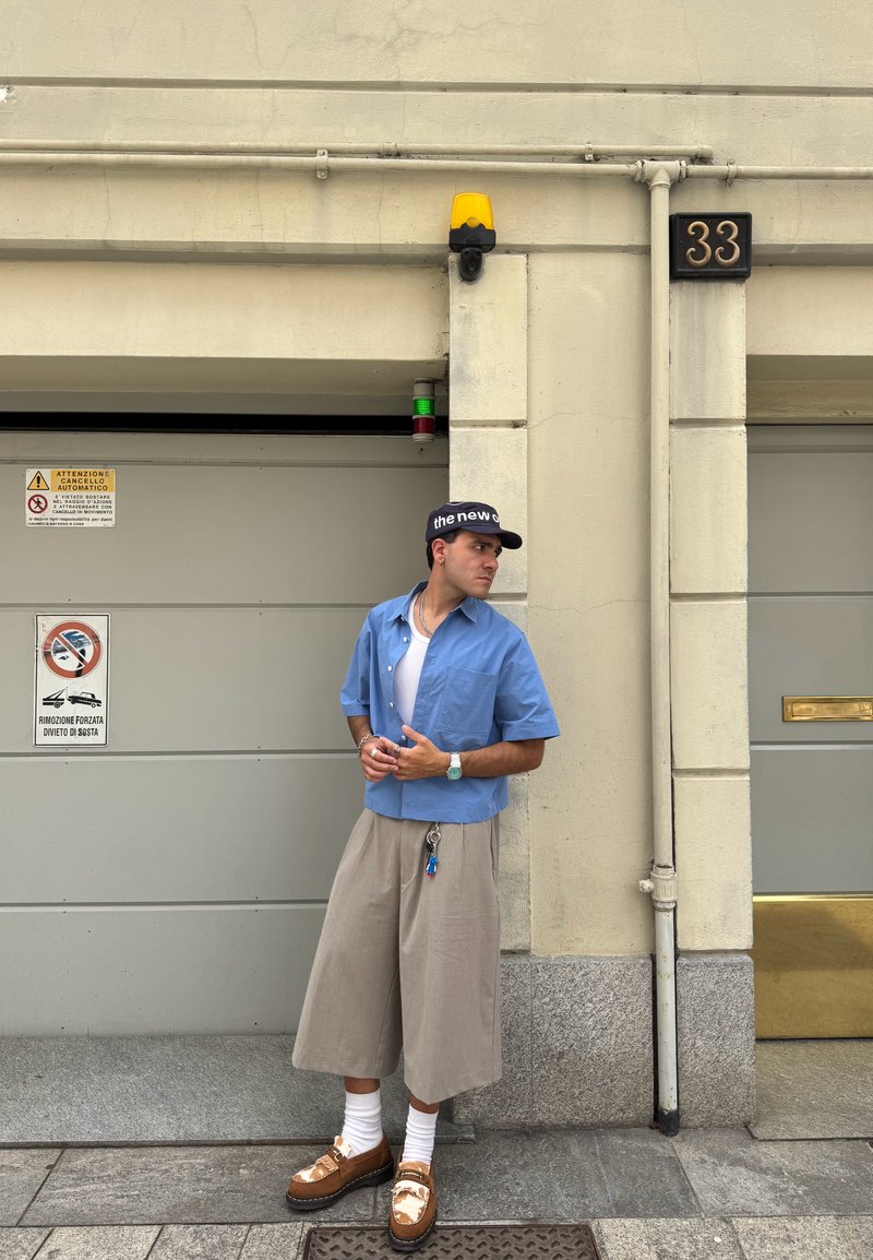 Chemise légère à manches courtes bleu clair, shorts beige amples, chaussures marron avec des chaussettes blanches, porte-clés attaché, capteurs muraux et panneaux d'avertissement visibles.