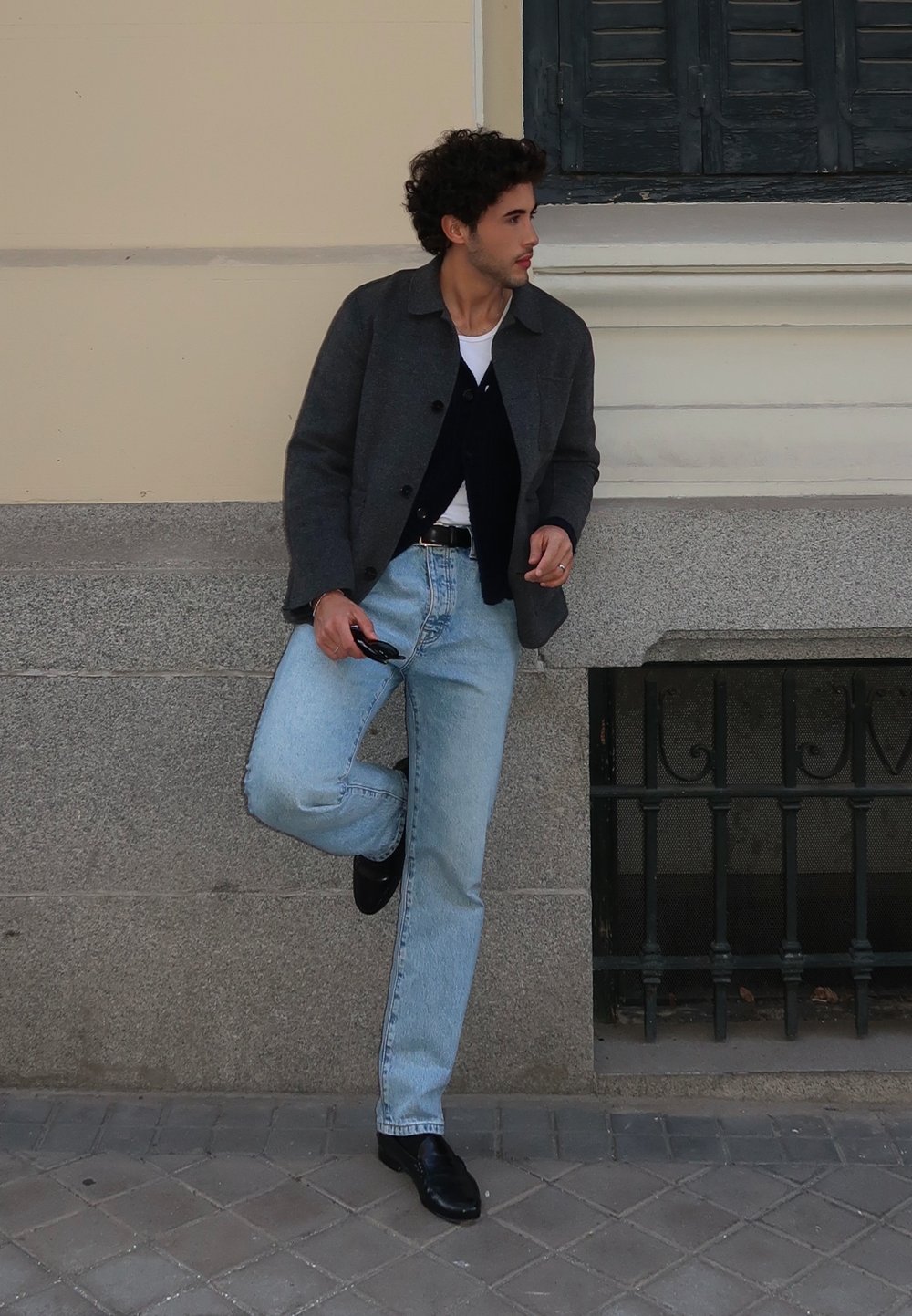 Blazer cinza de lã, cardigã azul-marinho, t-shirt branca, jeans azul-claro, sapatos de pele pretos. Encostado a uma superfície de pedra. Luz natural.