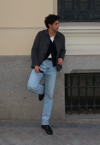 Blazer cinza de lã, cardigã azul-marinho, t-shirt branca, jeans azul-claro, sapatos de pele pretos. Encostado a uma superfície de pedra. Luz natural.