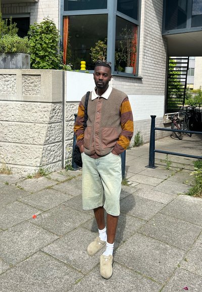 Persona vestida con una chaqueta de forro polar marrón y beige, con un cuello blanco, pantalones cortos de mezclilla claros, y zuecos beige, se encuentra de pie sobre una acera.