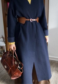 Manteau en laine bleu marine avec une taille cintrée par une ceinture, doté d'un col en V profond et de manches larges. Sac à main en cuir marron avec deux poignées, présentant une fermeture métallique brillante.