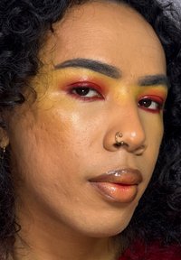 Gros plan sur un visage aux cheveux bouclés. Maquillage des yeux audacieux avec des tons jaunes et rouges. Lèvres brillantes dans des nuances de brun. Anneau de septum en or.