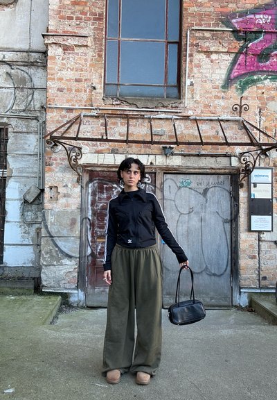 Chaqueta negra con cremallera y franjas blancas, pantalones anchos sueltos de color verde oliva, zapatillas beige y un pequeño bolso negro. Fondo de pared de ladrillos.