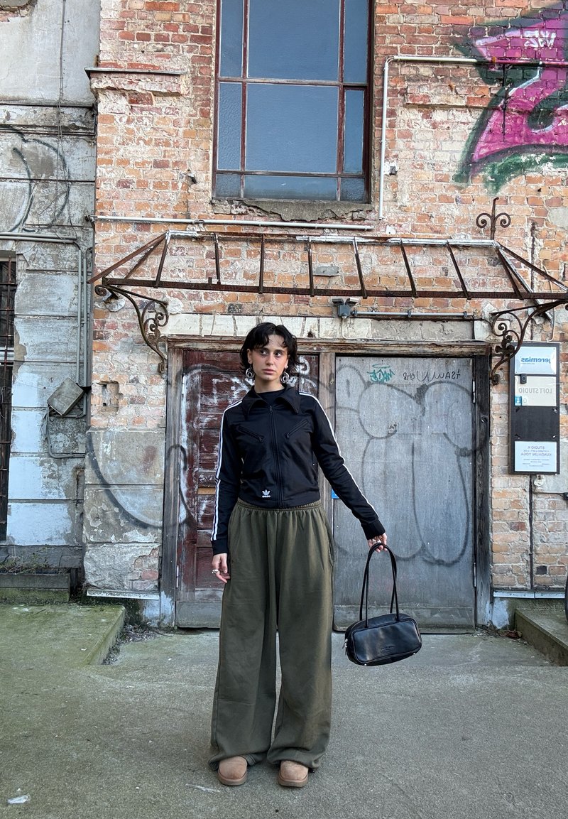 Casaco preto com fecho éclair e riscas brancas, calças largas verde-oliva folgadas, chinelos bege e uma pequena mala preta. Fundo de parede de tijolos.