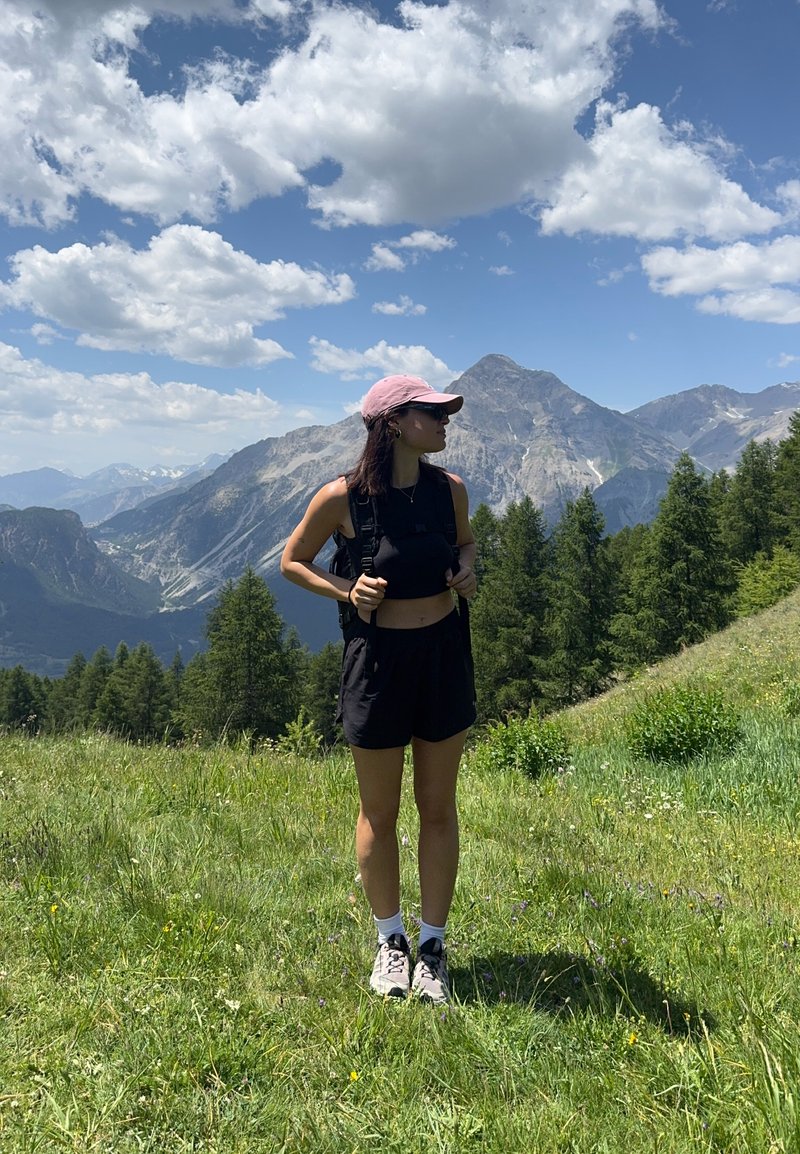 Femme portant un short noir et un haut avec une casquette rose et un sac à dos, debout sur une colline herbeuse avec des montagnes et des arbres sous un ciel partiellement nuageux.