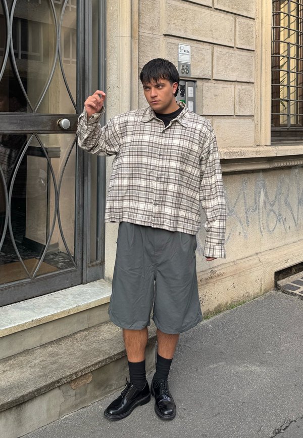 Camicia a quadri beige e marrone, design cropped; pantaloni corti grigi; scarpe nere; in piedi accanto a una porta con una cornice metallica decorativa.