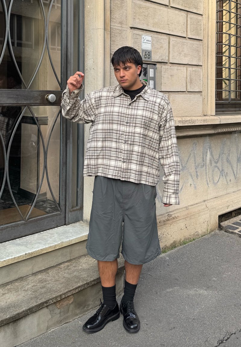 Camicia a quadri beige e marrone, design cropped; pantaloni corti grigi; scarpe nere; in piedi accanto a una porta con una cornice metallica decorativa.