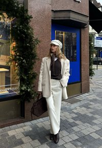 Manteau en laine crème, pull marron, pantalon blanc, chaussures en cuir marron et casquette claire. Sac à main marron et pavé texturé en arrière-plan.