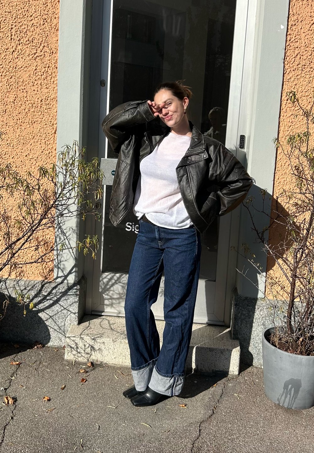 Czarna skórzana kurtka, biała koszula z długim rękawem, niebieskie dżinsy o szerokich nogawkach z jasnoróżowymi mankietami oraz czarne botki. Miejski plener na zewnątrz.