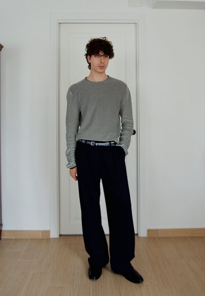 Joven de pie en el interior con cabello rizado, vestido con una camisa de manga larga a rayas blancas y negras, pantalones anchos negros y zapatos negros.