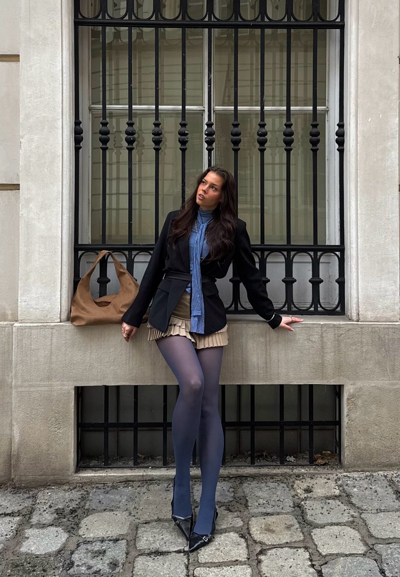 Femme en blazer noir, écharpe bleue, jupe plissée beige et collants bleu marine appuyée contre un rebord en pierre avec un sac marron à côté d'elle.