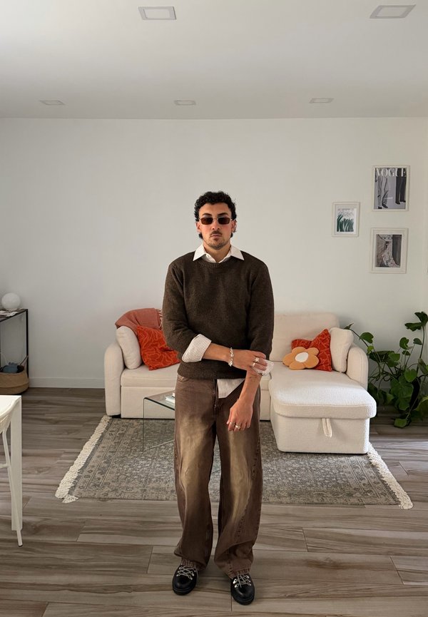 Homme aux cheveux bouclés et lunettes de soleil, portant un pull et un pantalon marron, debout dans un salon avec un canapé blanc et des plantes en arrière-plan.