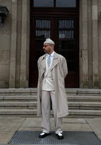Μπεζ παλτό με διπλό κούμπωμα πάνω από ένα ανοιχτόχρωμο γιλέκο, συνδυασμένο με φαρδύ παντελόνι. Γκρι σκουφάκι, στρογγυλά γυαλιά και μαύρα παπούτσια με λευκές λεπτομέρειες.