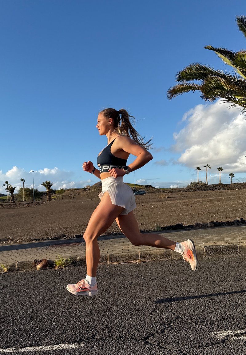 Athlète courant sur un trottoir, portant un soutiens-gorge de sport noir, un short blanc et des baskets roses avec des chaussettes blanches, sur fond de ciel bleu clair.