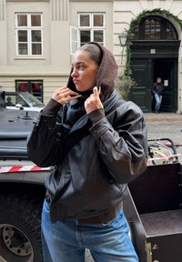 Jeune personne ajustant une capuche marron sur la tête, portant une veste en cuir noire et un jean, debout sur une rue pavée près d'un véhicule et d'une entrée de bâtiment.