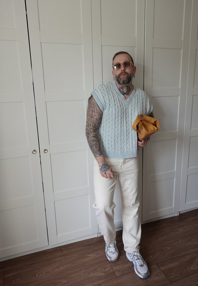 Hombre con tatuajes y gafas de sol, que lleva un suéter sin mangas de punto azul, pantalones blancos, zapatillas deportivas, sosteniendo una bolsa marrón plegada en interiores.