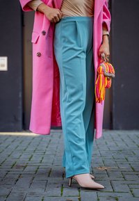 Ένα μακρύ ροζ παλτό με μεγάλα κουμπιά πάνω από τιρκουάζ παντελόνι, σε συνδυασμό με μπεζ ψηλοτάκουνα παπούτσια και μία πολύχρωμη, υφασμάτινη τσάντα.