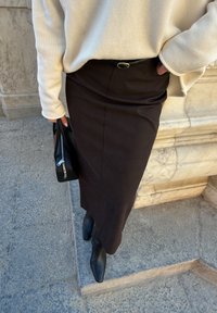 Personne portant un pull crème, ceinture noire, jupe midi marron foncé, bottes à talons noires, tenant un sac à main noir, debout sur un pavé en pierre près d'un mur.