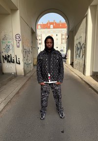 Casaco de ganga preto com um padrão repetido, combinado com calças pretas estampadas. O conjunto inclui uma camisa branca por baixo e ténis pretos.