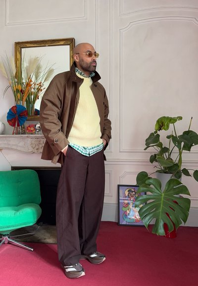 Hombre con gafas de sol, chaqueta marrón, suéter amarillo, camisa a cuadros verde, pantalones anchos marrones, de pie en el interior junto a una silla verde, una planta grande y arte enmarcado.