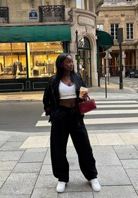 Kvinna i svarta byxor och vit crop top som håller en iskall kaffe och en röd väska, stående på en gata i Paris nära ett övergångsställe och en butik.