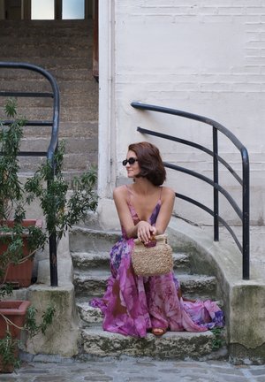 Vestido morado fluido con flores y tirantes finos, bolso de fibra beige, sentado en escaleras de piedra, rodeado de plantas en macetas y una barandilla.