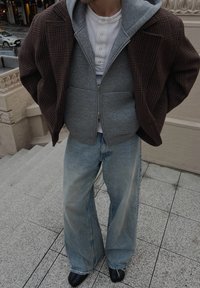 Personne portant des vêtements superposés : chemise henley blanche, sweat à capuche gris zippé, blazer à carreaux marron, jean bleu clair ample et chaussures noires fendue à l'avant.