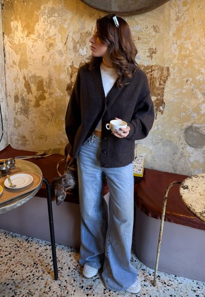 Mujer con cárdigan oscuro y vaqueros claros sosteniendo una taza de café, de pie en una cafetería rústica con paredes texturizadas y suelo de terrazo.