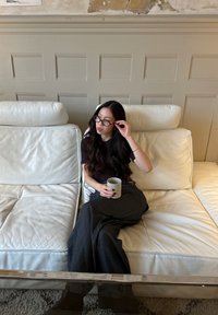 Divano in pelle bianca con due cuscini; una persona con maglia scura e pantaloni a righe tiene una tazza chiara, con capelli lunghi e occhiali.