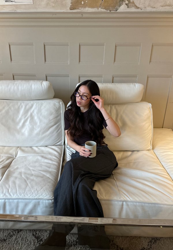 Sofá de couro branco com duas almofadas; uma pessoa com uma blusa escura e calças riscas segura uma caneca de cor clara, com cabelo comprido e óculos.