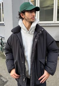 Geacă neagră matlasată cu guler mare, fermoare și capse, purtată peste un hanorac gri. Șapcă verde cu litere albe.