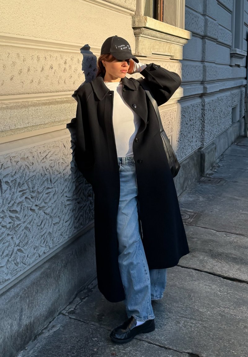 Persona che indossa un lungo cappotto nero, camicia bianca, jeans blu, cappello nero e scarpe nere, in piedi contro un muro di pietra lavorata all'aperto.