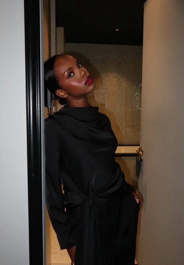 Frau mit dunkler Haut und rotem Lippenstift lehnt an einem Türrahmen und trägt eine schwarze Bluse mit hohem Kragen sowie passende Hosen in einem schwach beleuchteten Raum.