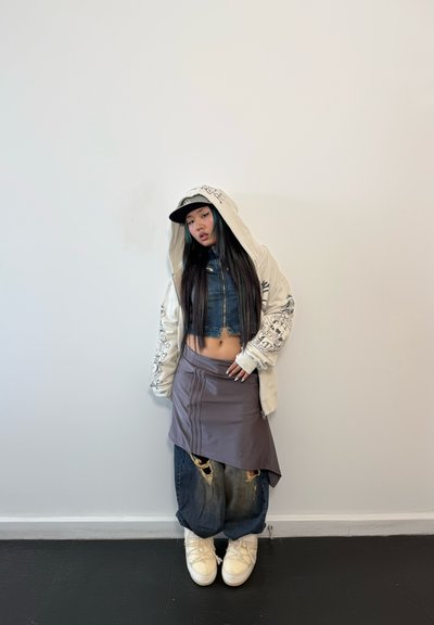 Sudadera de gran tamaño color crema con detalles gráficos, chaqueta de mezclilla corta, falda asimétrica gris, jeans oscuros desgastados y zapatillas blancas chunky.