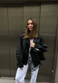 Γυναίκα με μαλλιά μέχρι τον ώμο φοράει μαύρο δερμάτινο μπουφάν πάνω από λευκό παντελόνι και διαφανές τοπ με ανθισμένο σχέδιο, στέκεται μπροστά σε μεταλλικές πόρτες.