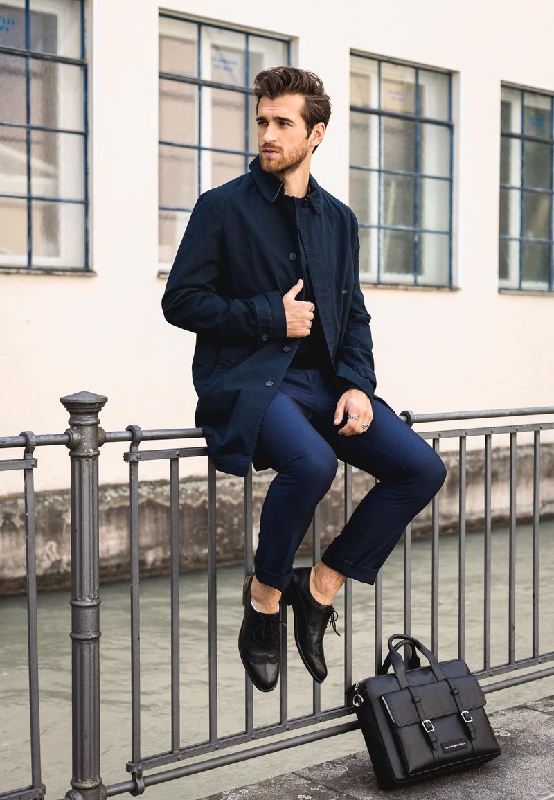 Zwarte jas, marineblauwe broek, zwarte leren schoenen; zittend op een leuning naast een zwarte tas met zilveren hardware en dubbele gesps accenten.