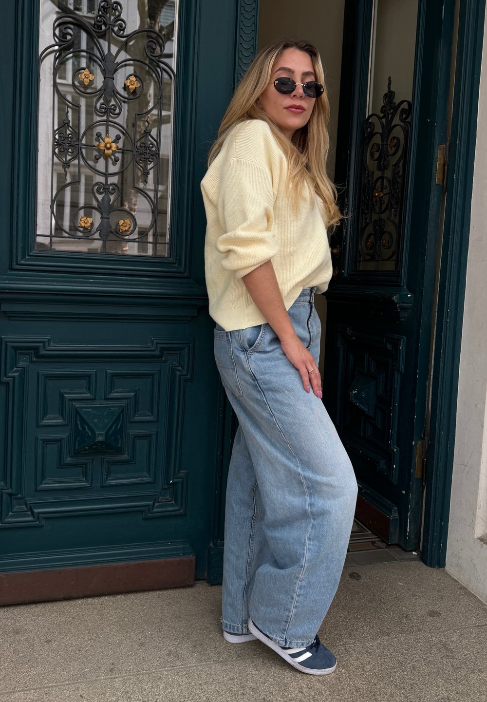 Sweater oversized amarelo claro, jeans de ganga azul claro de cintura alta e ténis azul-marinho, de pé contra uma porta ornamentada verde escura.