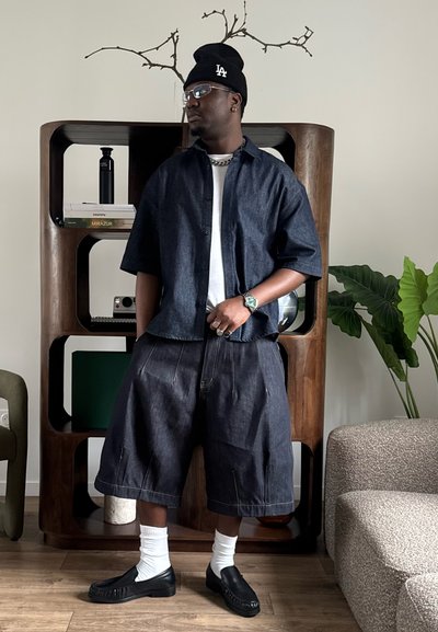 Hombre con camisa vaquera oscura, pantalones cortos holgados, calcetines blancos, mocasines negros y gorro negro, de pie en el interior cerca de una estantería de madera y una planta.