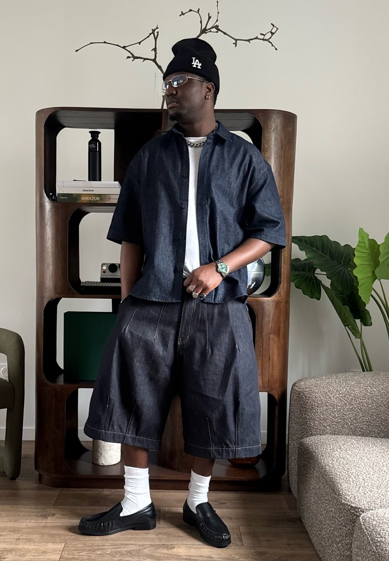 Homme portant une chemise en jean foncé, un short ample, des chaussettes blanches, des mocassins noirs et un bonnet noir, debout à l’intérieur près d’une étagère en bois et d’une plante.